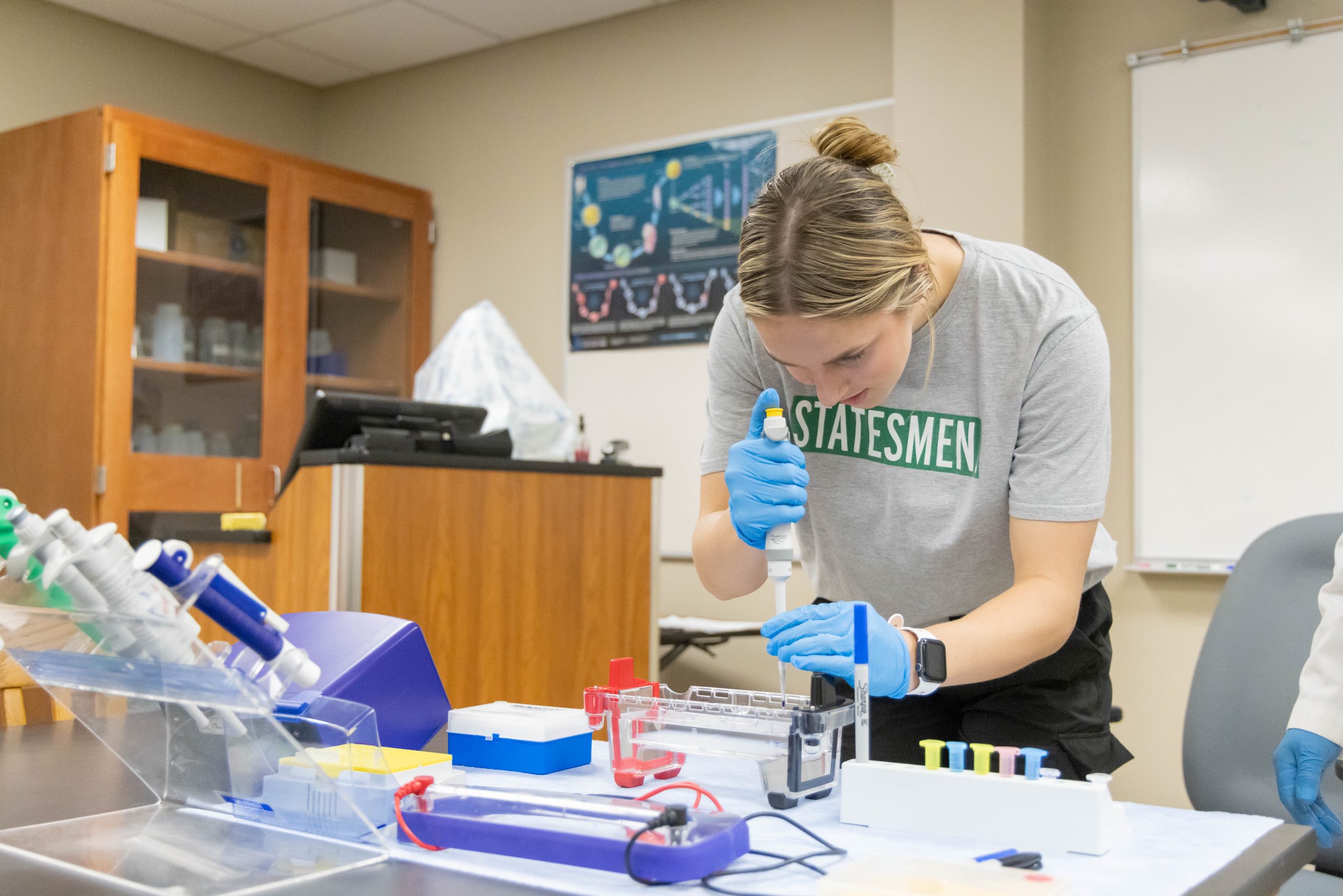 Delta State student wearing a "Statesmen" shirt conducting a lab experiment using pipettes and lab equipment in a science classroom.