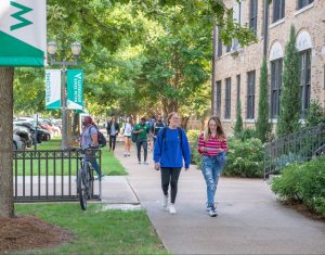 Delta State University students walking and biking along a tree-lined campus sidewalk near academic buildings, with welcome banners displayed.