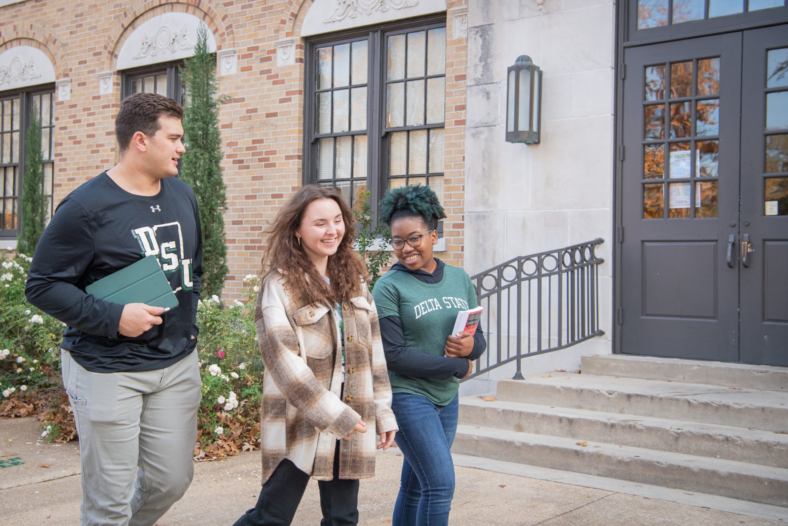 Three Delta State University students walking and smiling outside a campus building, holding notebooks and wearing DSU apparel.