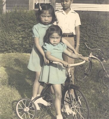 Brother and two sisters posing near tricycle. Color.