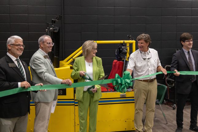 university leaders and a government official preparing to cut a green ribbon in front of a new aircraft flight simulator.