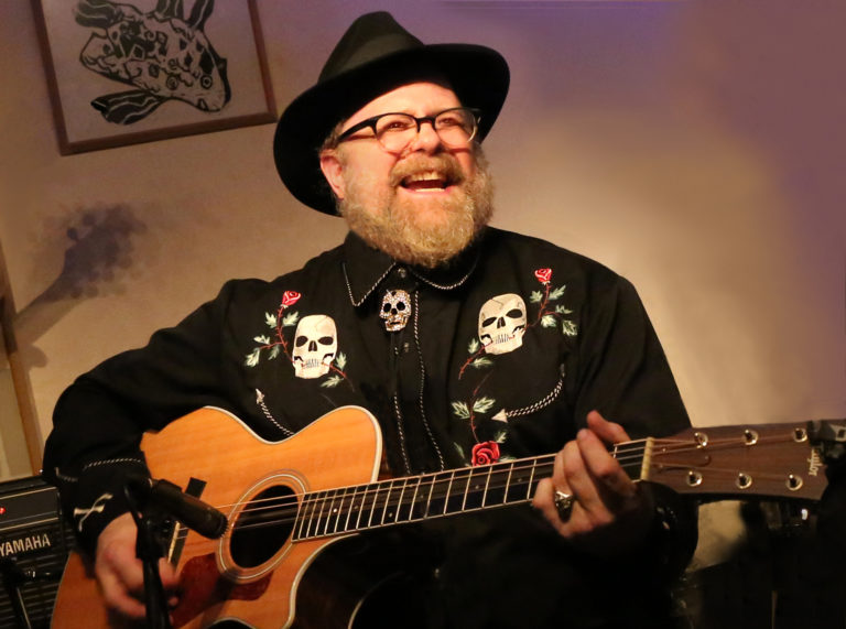 a bearded man in a black hat and shirt playing guitar.