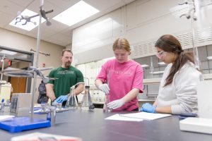 Students conducting a chemistry experiment with guidance from a faculty member in a laboratory.