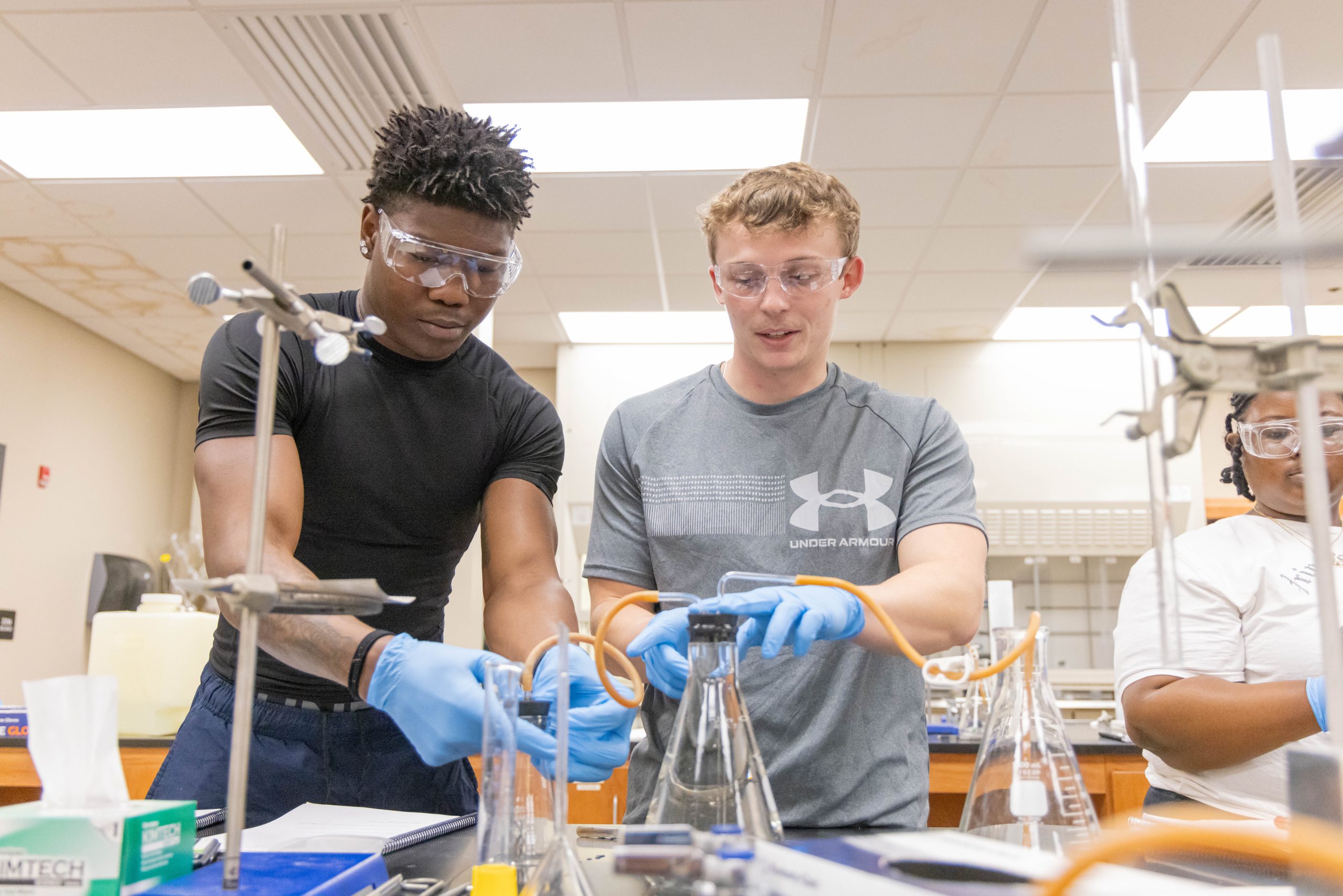 Students wearing protective eyewear and gloves working together on a chemistry experiment.