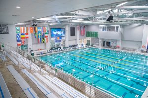 Indoor aquatic center with an Olympic-sized swimming pool, spectator seating, and international flags.