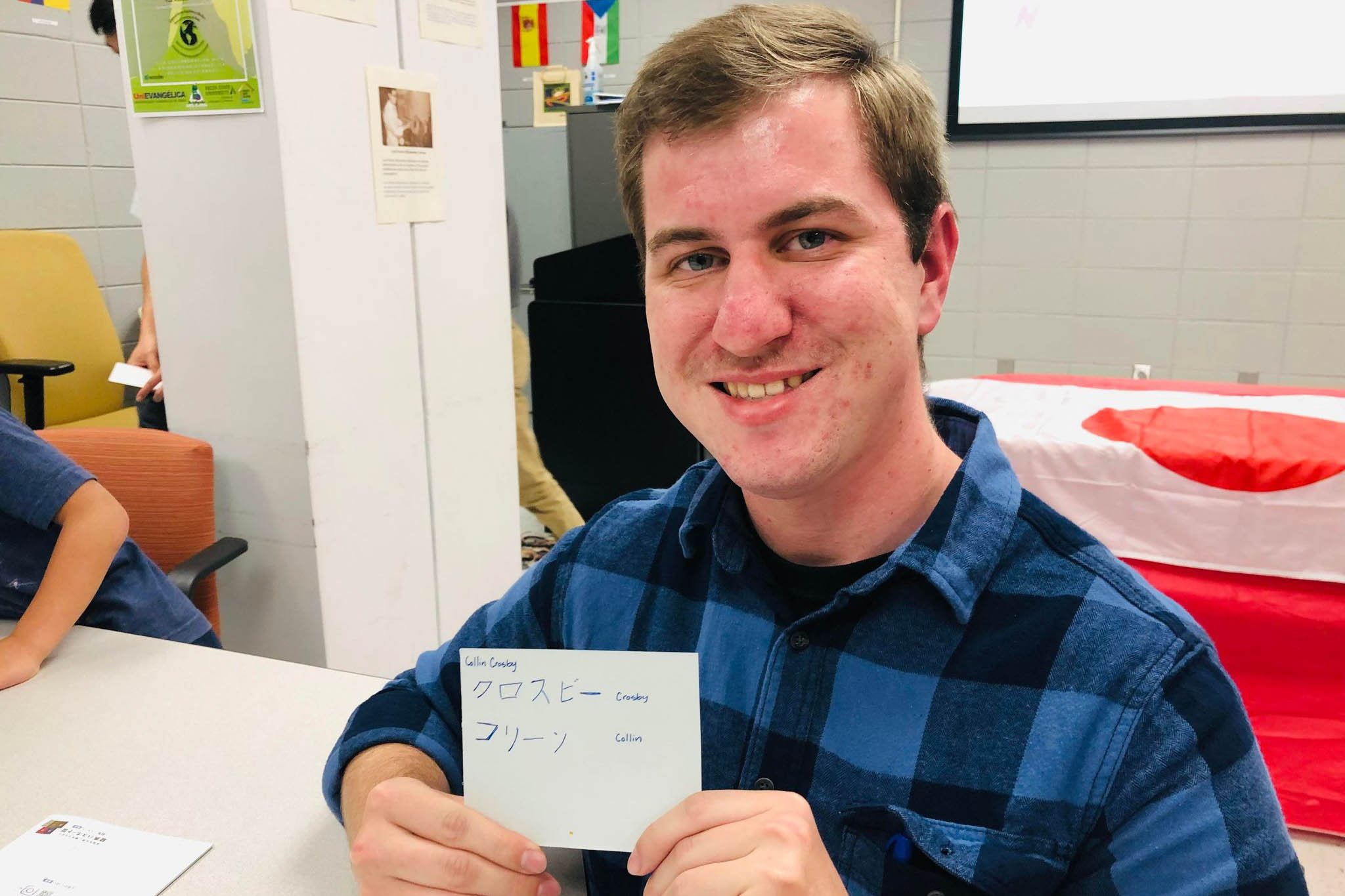 Collin Crosby smiling and holding a card with Japanese writing, seated at a table with an international flag behind him in the background.
