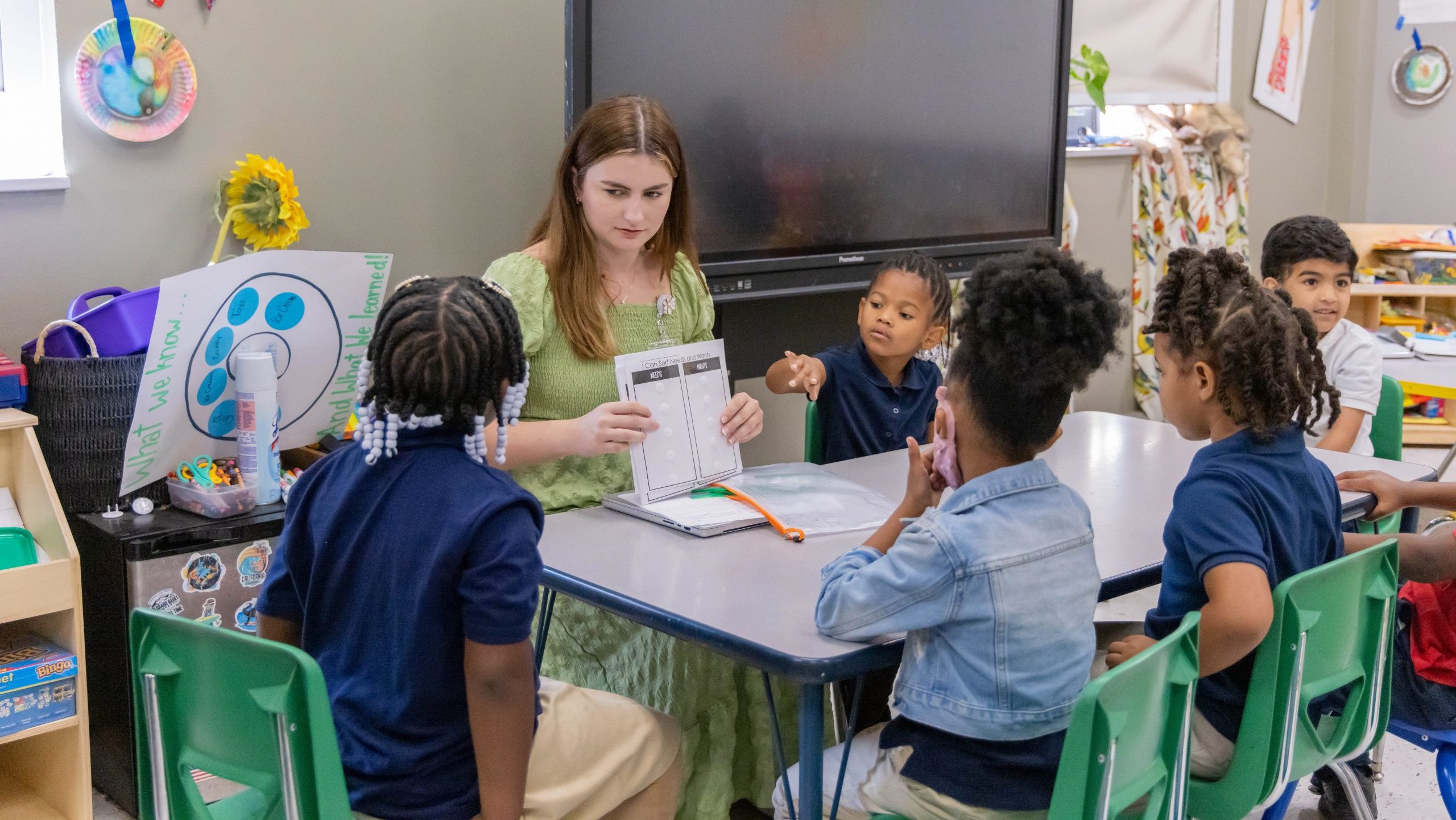 Teacher sitting at a small table with a group of young children, holding a worksheet during a classroom activity.
