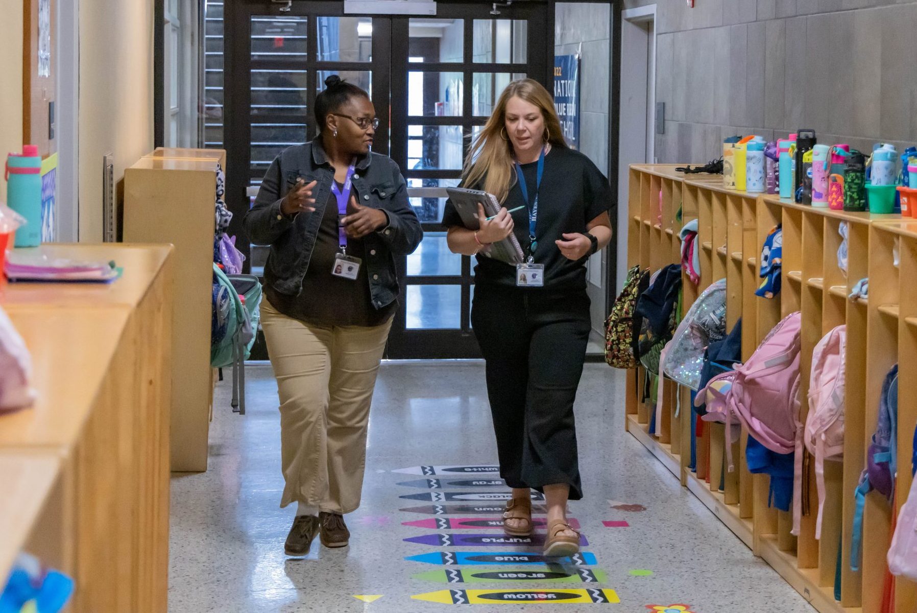 Two women talking while walking down an elementary school hallway that has cubbies filled with student belongings lining the walls.