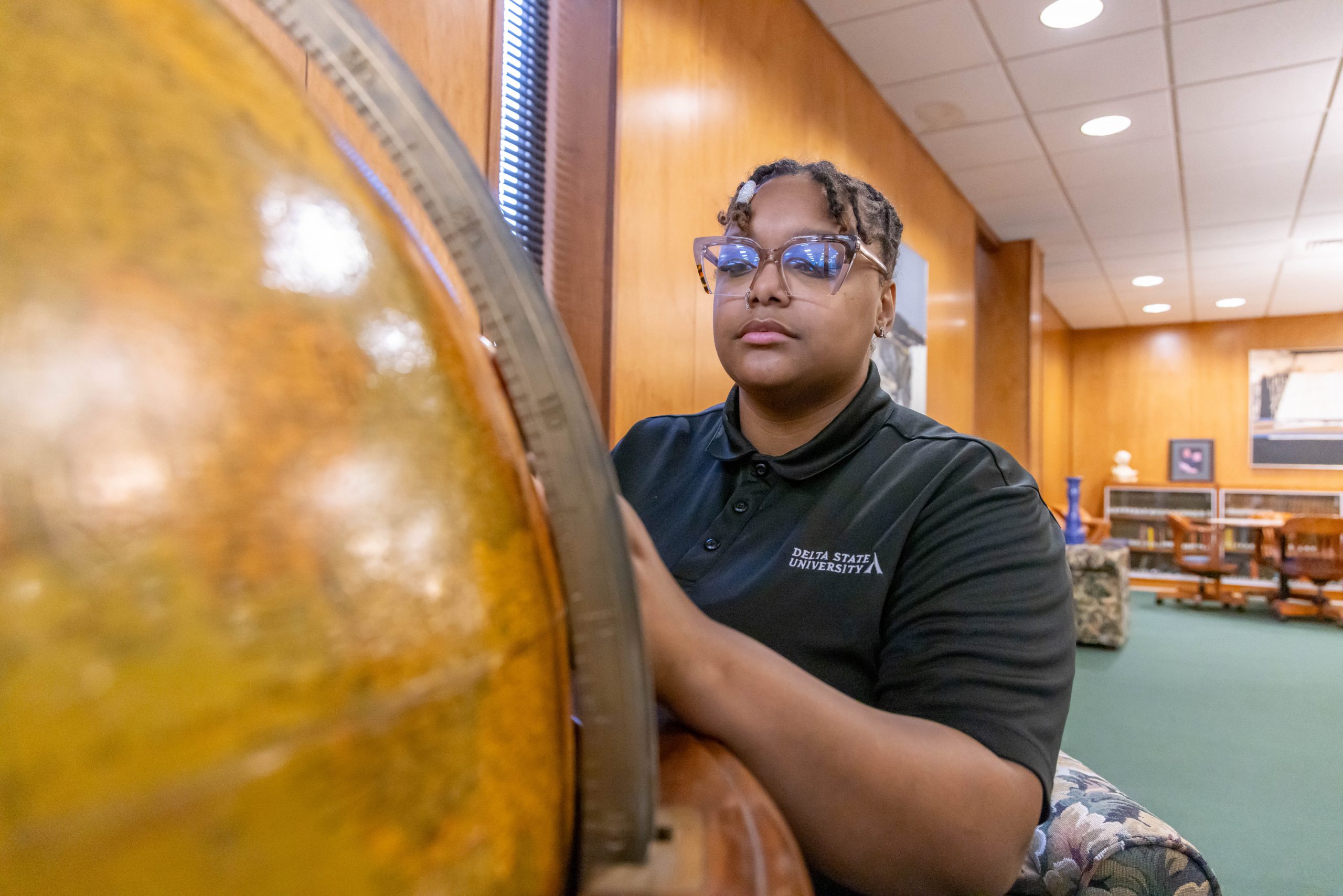 A female student wearing a Delta State University polo shirt and glasses closely examining a large globe in a well-lit library study space with bookshelves and seating areas.