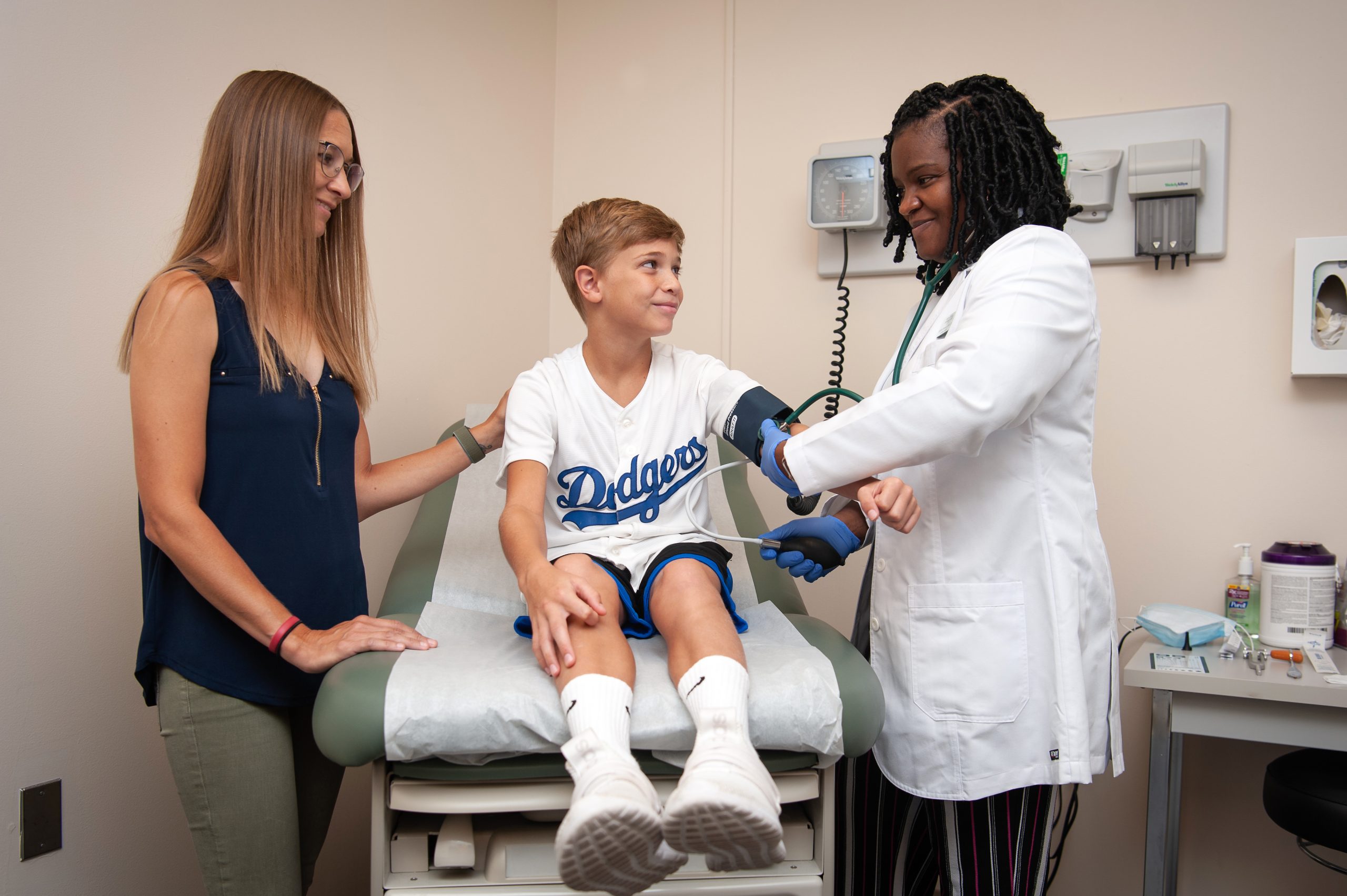 Healthcare professional checking a child's blood pressure while a parent looks on in a medical exam room.